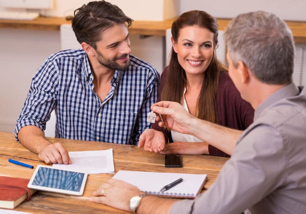 Young happy couple receiving house keys from real estate agent. Giving keys of new house to young couple. Smiling couple signing financial contract for mortgage.